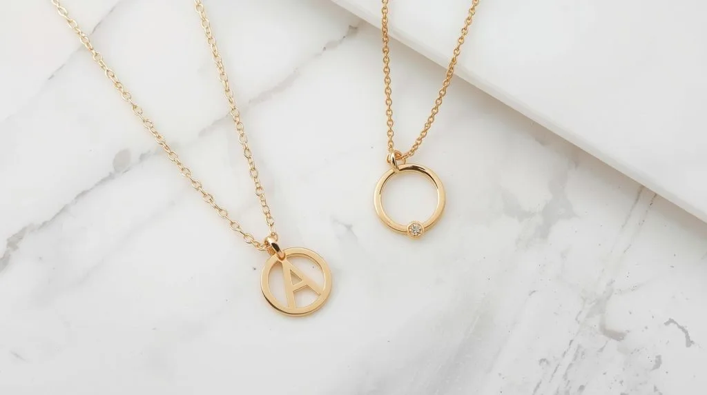 flat lay of minimal gold jewellery — thin initial pendant, small stud earring and delicate chain — on a marble surface. soft, clean, editorial.